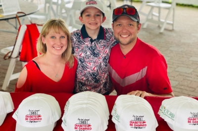 Brent posing with son and wife at the annual golf event in front of a table of hats that say: 